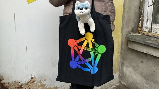 Torba z logo Fediwersum, z której wystaje maskotka pieska. W tle Łódź.